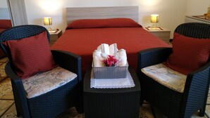 Habitación de hotel con cama, dos sillones, una mesita con un cesto de artículos de aseo y un cuadro en la pared.
