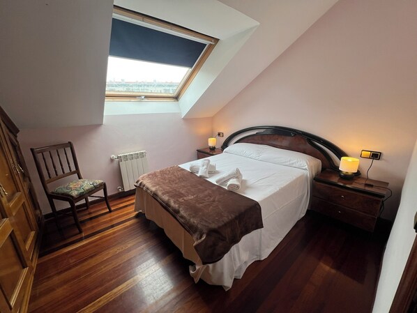 A bedroom with a sloped ceiling, a skylight, a wooden floor, a bed with white linens, a chair, a bedside table with a lamp, and a radiator.