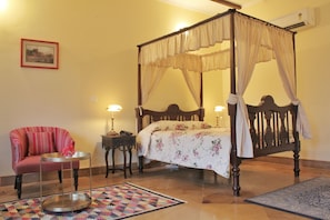 Ein Bett mit Baldachin, mit floralem Überwurf, ein roter, gepolsterter Sessel, ein kleiner runder Tisch mit Tablett und ein gemustertes Teppich auf Fliesen.