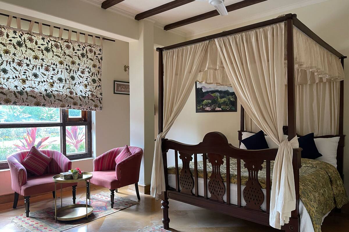 A four-poster bed with a canopy, a chair with a patterned cushion, a small table with a tray, and a window with a decorative curtain.