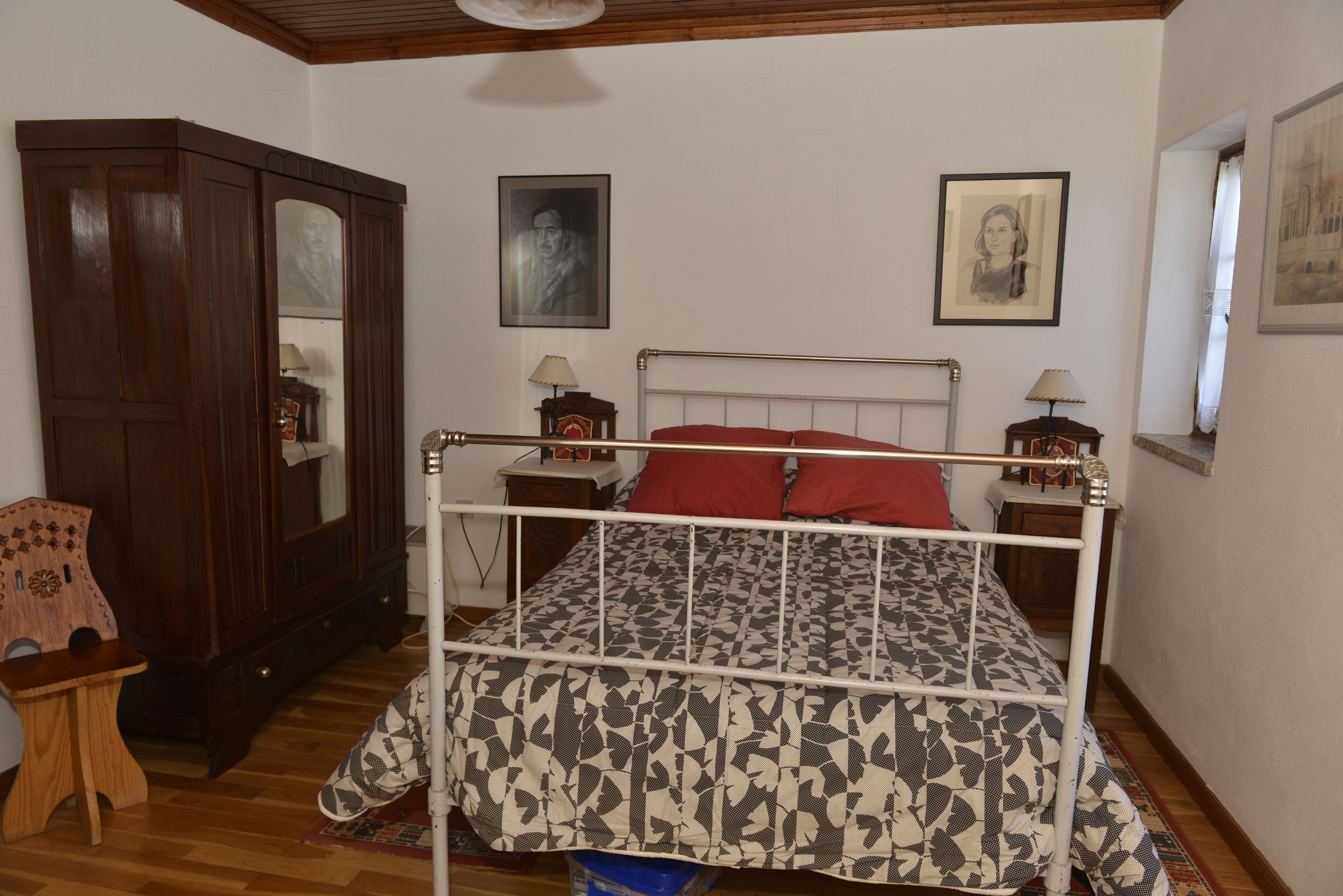 A bedroom with a bed, wooden wardrobe, and two framed portraits on the wall.