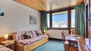 Ein gemütliches Zimmer mit Holzbalkendecke, einer Couch mit gemusterten Kissen, einem Esstisch mit Stühlen und einem Fenster mit Vorhängen.