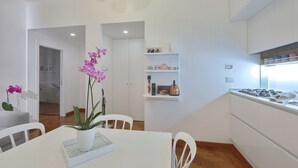 Una cucina moderna con mobili bianchi, un piano cottura a gas e un ripiano di lavoro bianco. C'è un'area pranzo con un tavolo e sedie bianche e una pianta di orchidea in vaso.