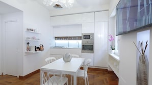 Una cucina moderna con mobili bianchi, un tavolo da pranzo con sedie, una TV a parete e un vaso con fiori sul davanzale.
