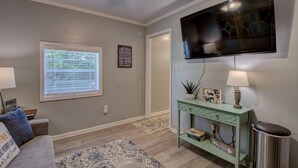 Un salon avec un téléviseur à écran plat fixé au mur, une console verte sur laquelle est posée une lampe, et une fenêtre équipée de stores.