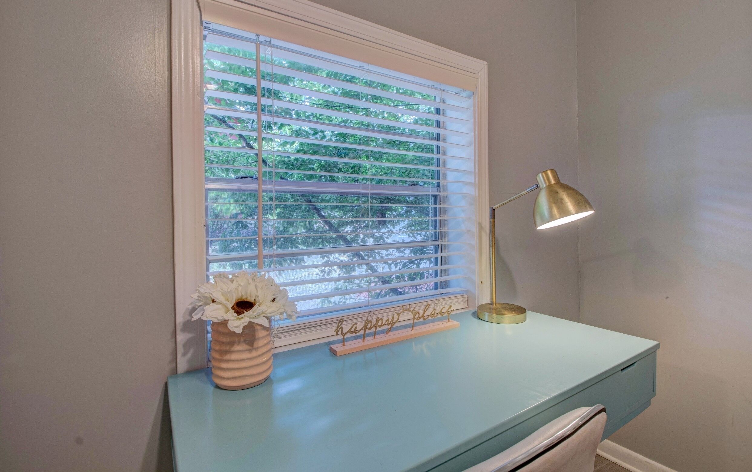 A room with a window covered by blinds, a desk with a lamp, and a vase with flowers.