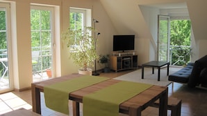 A spacious living area with a wooden table, a sofa, and a television on a stand. There are large windows providing natural light and a view of greenery outside. - Ferienwohnung Buhl (Regensburg)