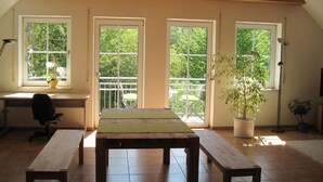 A room with a wooden table, two benches, a desk with a chair, and three windows with a view of greenery. - Ferienwohnung Buhl (Regensburg)