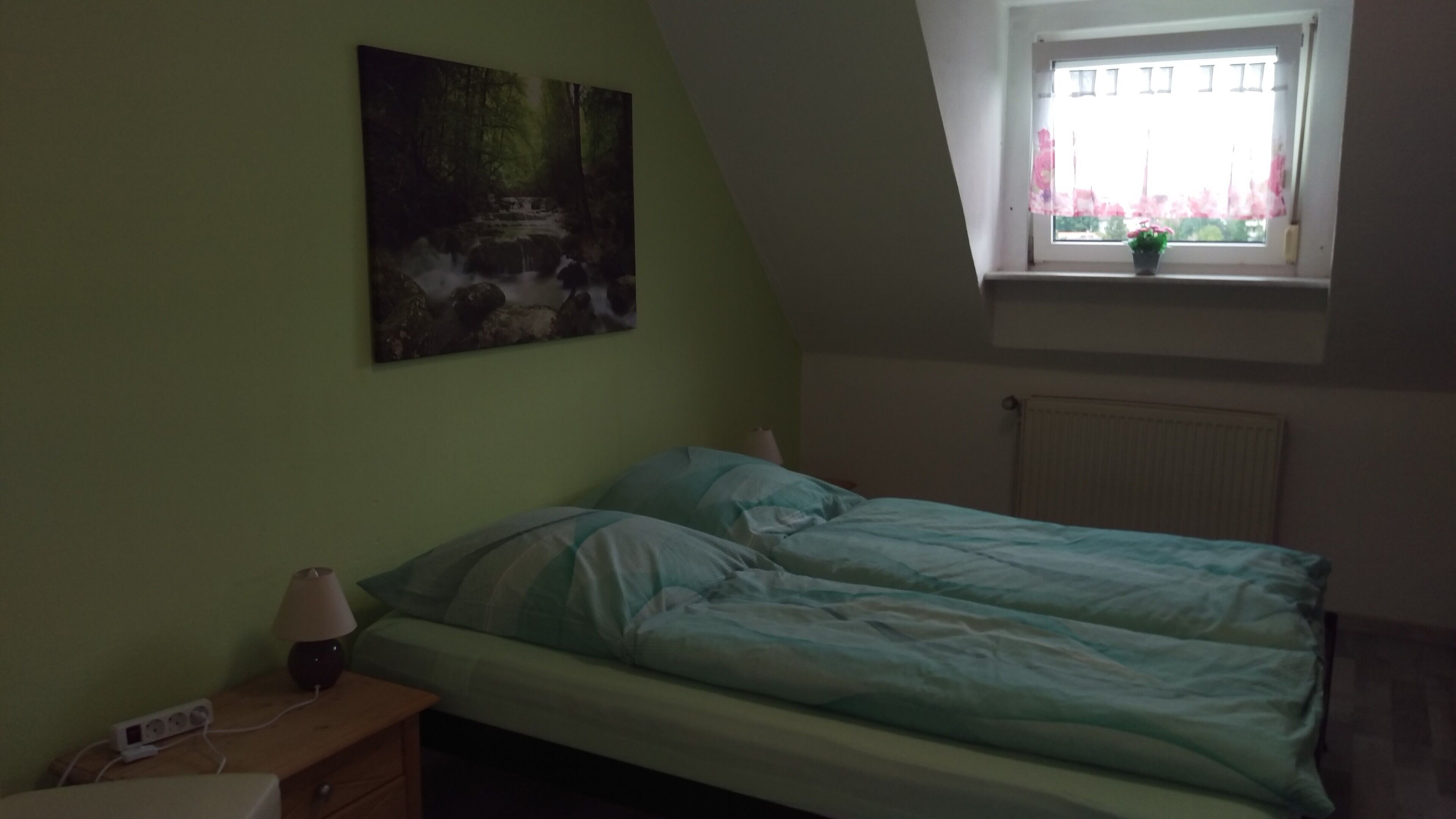 Quarto com cama, mesa de cabeceira, luminária, régua de tomadas, janela com vista para um edifício e radiador.