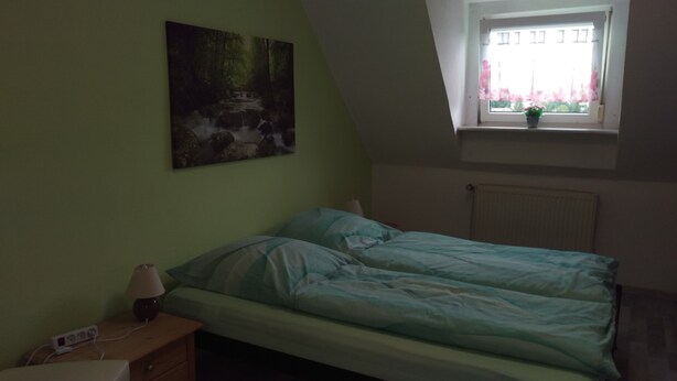 A bedroom with a bed, a nightstand, a lamp, a power strip, a window with a view of a building, and a radiator. - Ferienwohnung Fusi (Amberg)
