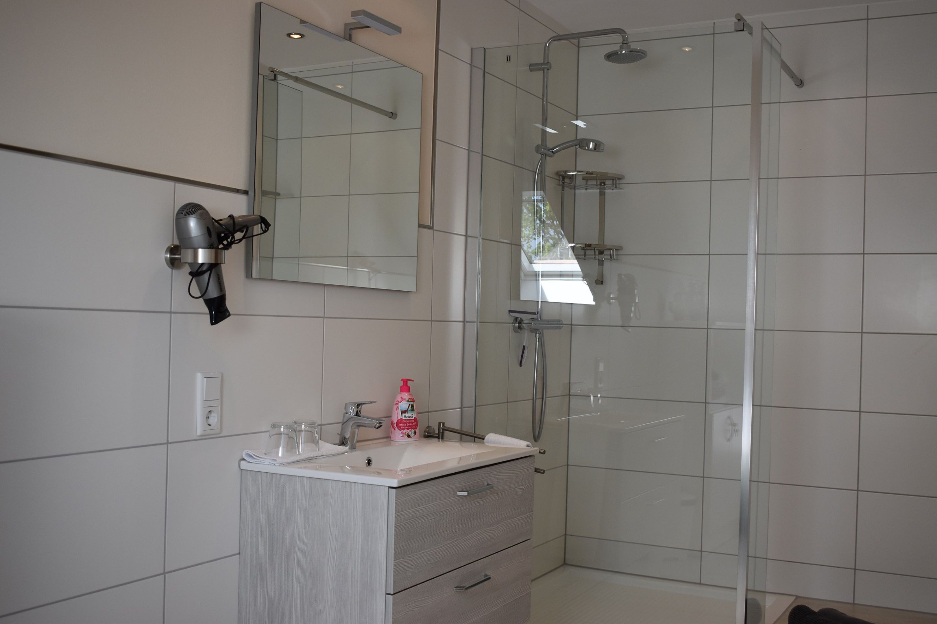 Une salle de bain moderne dotée d’une douche à parois de verre, d’un sèche-cheveux fixé au mur et d’un meuble-lavabo avec un miroir.