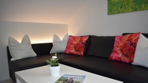 En moderne stue med en sort sofa, hvide puder, et hvidt sofabord med et magasin og en lille urtepotte.