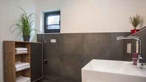A modern bathroom with a large bathtub, a wooden shelf with towels, a potted plant, and a window. - Ferienwohnung Happyliving (Winterberg)