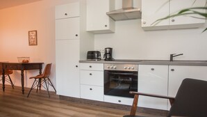 A compact kitchen with white cabinets, a built-in oven, a coffee maker, and a dining area with a table and chairs. - Ferienwohnung Happyliving (Winterberg)
