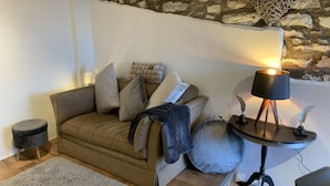 Una sala de estar acogedora con un sofá marrón, varios cojines, una mesita redonda con una lámpara y una pared de piedra.