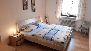 A bedroom with a bed, nightstand, chair, and a window with curtains. - Ferienwohnung Fürst (Mauth)