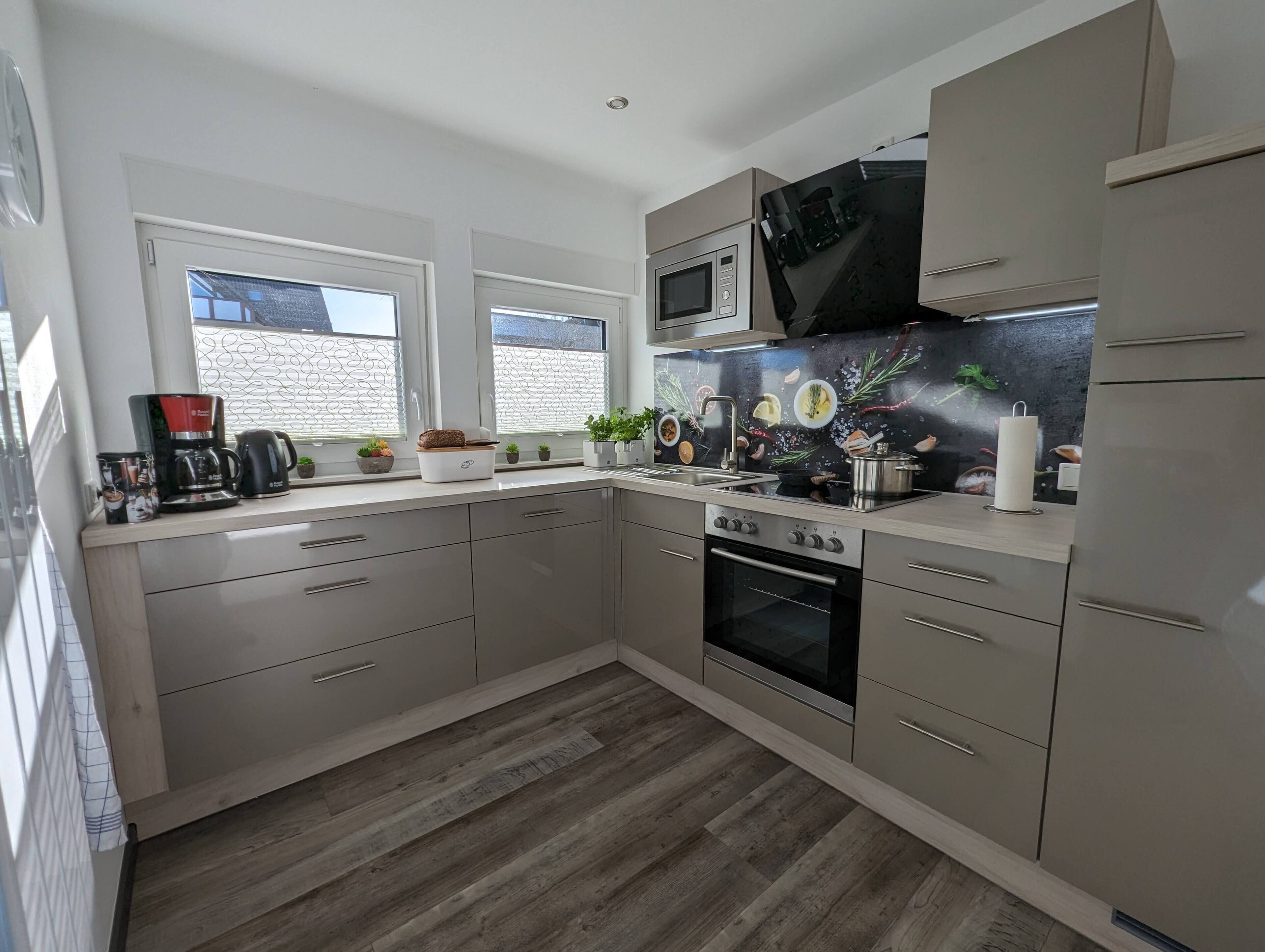 A modern kitchen with a built-in oven, microwave, and stainless steel appliances. There are two windows with decorative grills, a sink, and a countertop with various kitchen items.