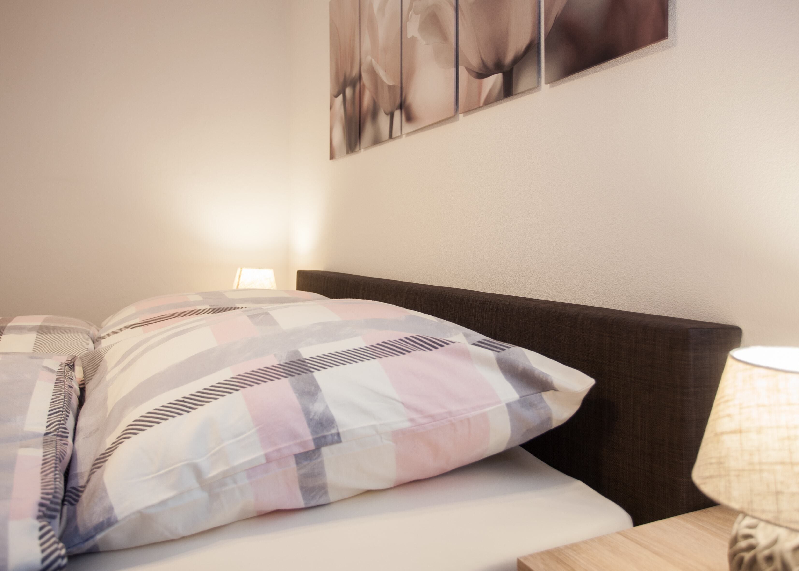 A neatly made bed with a checkered pillow and a headboard. There is a wall-mounted lamp and a framed picture above the bed.