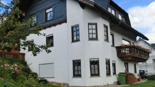 Une maison à deux étages, avec une façade blanche et un toit foncé, dotée de nombreuses fenêtres et d’un balcon agrémenté de plantes.