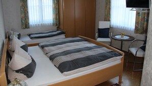 Habitación de hotel con una cama grande, dos camas más pequeñas, un armario, un televisor, una mesita con una silla y cortinas.