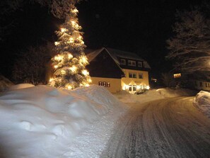 A snow-covered landscape with a lit Christmas tree and a house with a visible sign.