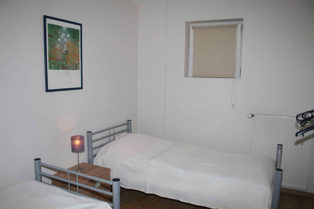 Uma cama de solteiro com estrutura de metal, uma mesinha de cabeceira com abajur, um quadro na parede e uma janela com cortina.