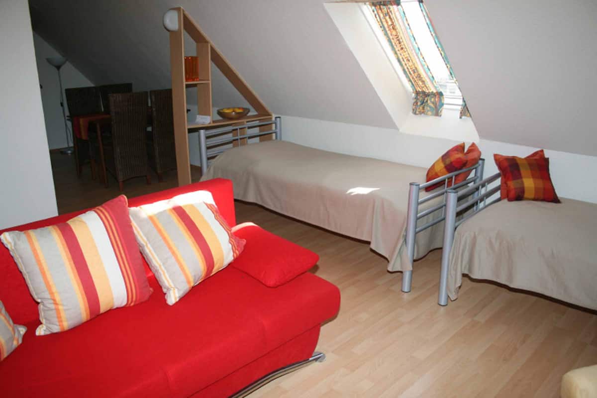 Quarto compacto com sofá vermelho, duas camas simples, escada de madeira e pequena área de refeições.