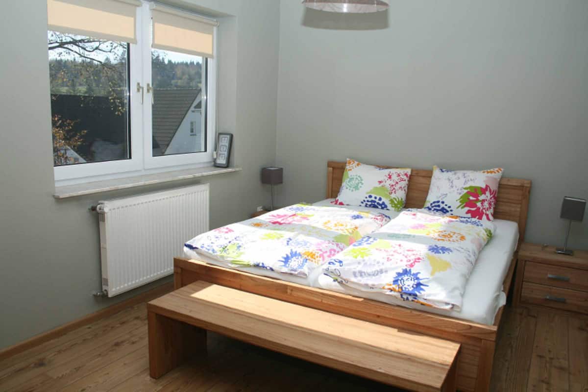 Quarto com cama de madeira, janela com vista para árvores, mesa de cabeceira com abajur e radiador.