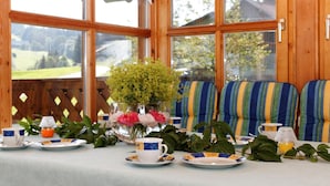 A dining area with a table set for tea, a vase of flowers, and a view of a green landscape through large windows. - Ferienhof Degenhart (Mauth)