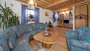 A living room with a floral sofa, a wooden coffee table, and a kitchen area with wooden cabinets and a TV. - Ferienhof Degenhart (Mauth)