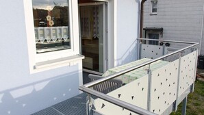 Un balcón con baranda de metal, una ventana con arreglo floral y una puerta que da a un espacio interior.