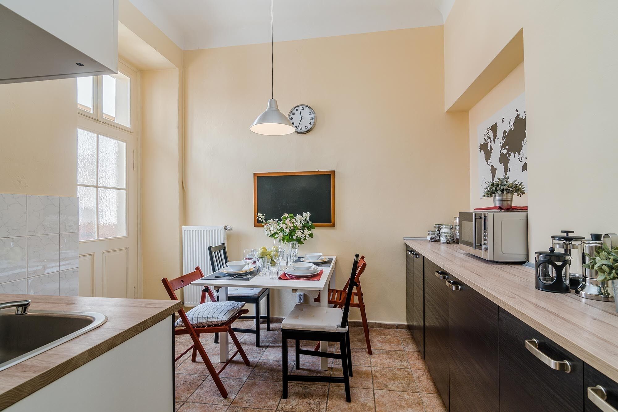 A kitchen with a dining table, chairs, a hanging light, a clock, a world map, and kitchen appliances.
