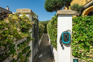 A narrow alleyway with a wall-mounted electric vehicle charging station, surrounded by lush greenery and a clear blue sky.