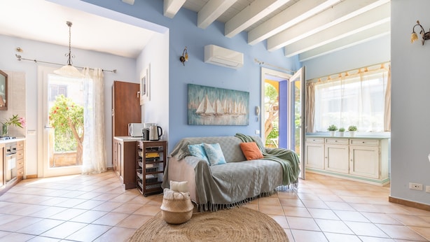 A spacious living area with a large window, a sofa, a coffee table, and a painting on the wall. - Holiday Home "Junco" with Garden, Loggia and Wi-Fi (Villasimius)