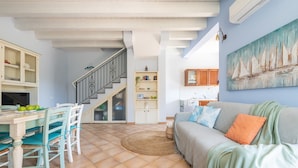 A living room with a sofa, a dining table with chairs, a staircase, and a kitchen area. - Holiday Home "Junco" with Garden, Loggia and Wi-Fi (Villasimius)