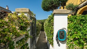 A narrow alleyway with a wall-mounted electric vehicle charging station, surrounded by lush greenery and a clear blue sky. - Holiday Home "Alloggio Nettuno" with Garden, Loggia and Wi-Fi (Villasimius)