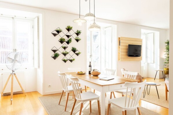 Un espace de repas lumineux, avec une table en bois et des chaises blanches, un téléviseur fixé au mur et des œuvres d’art géométriques accrochées au mur.