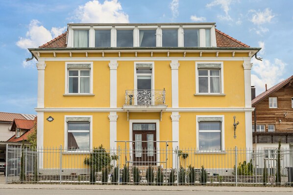 A two-story yellow building with white trim, a balcony, and a gated entrance. - Stylish Holiday Apartment "Tuna" in Old Building Villa, with Mountain View (Sigmarszell)
