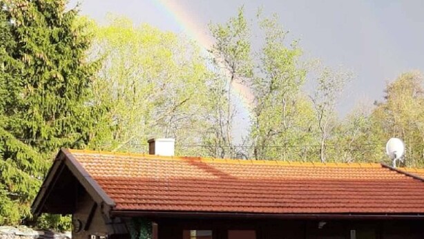 A wooden cabin with a red roof, surrounded by a white picket fence, and a rainbow in the background. - Gerda's Ferienhäuser (Eppenschlag)