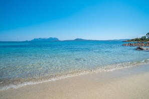 A beach with clear water, a distant sailboat, and rocky terrain.