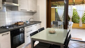 Une cuisine équipée d’une cuisinière à gaz, d’un four et de meubles blancs. On y trouve une table à manger avec des chaises et un plateau en verre.