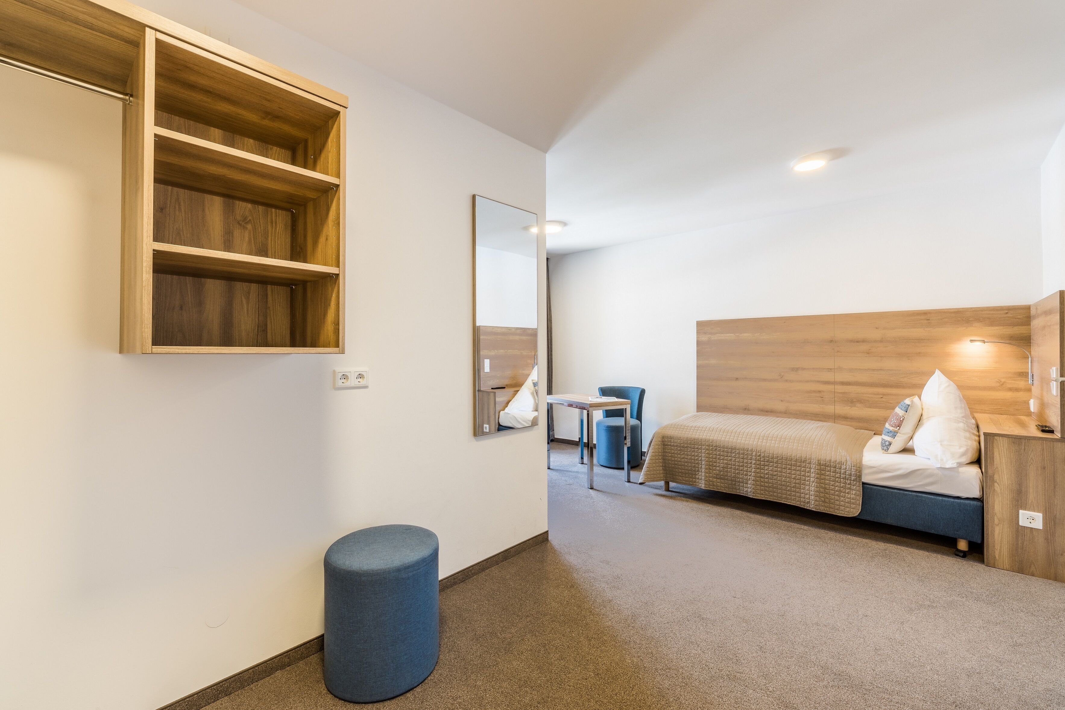 A compact hotel room with a bed, a small desk, a chair, a round ottoman, a wooden shelf unit, and a mirror.