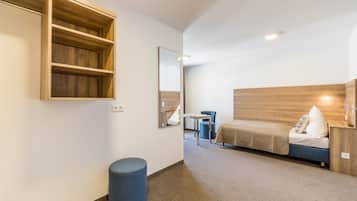 A compact hotel room with a bed, a small desk, a chair, a round ottoman, a wooden shelf unit, and a mirror.