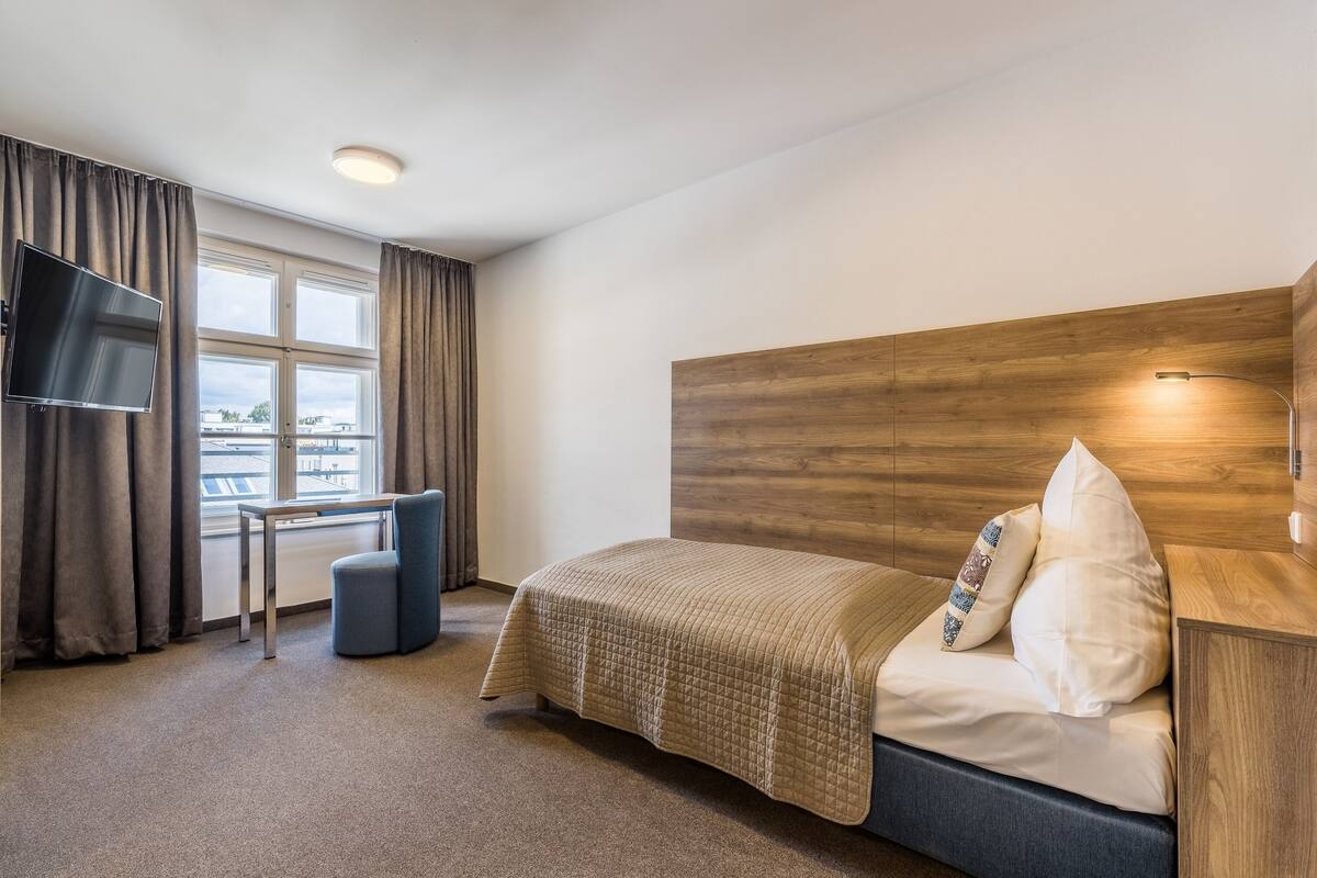 Habitación de hotel con cama, escritorio, silla, televisión y ventana con cortinas.