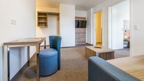 Habitació moderna i ben il·luminada amb escriptori, cadira blava i taula petita. Hi ha un armari de fusta i una porta que condueix a una altra habitació.