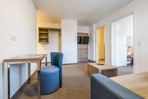 Una habitación moderna y luminosa con un escritorio, una silla azul y una pequeña mesa. Hay un armario de madera y una puerta que da a otra habitación.
