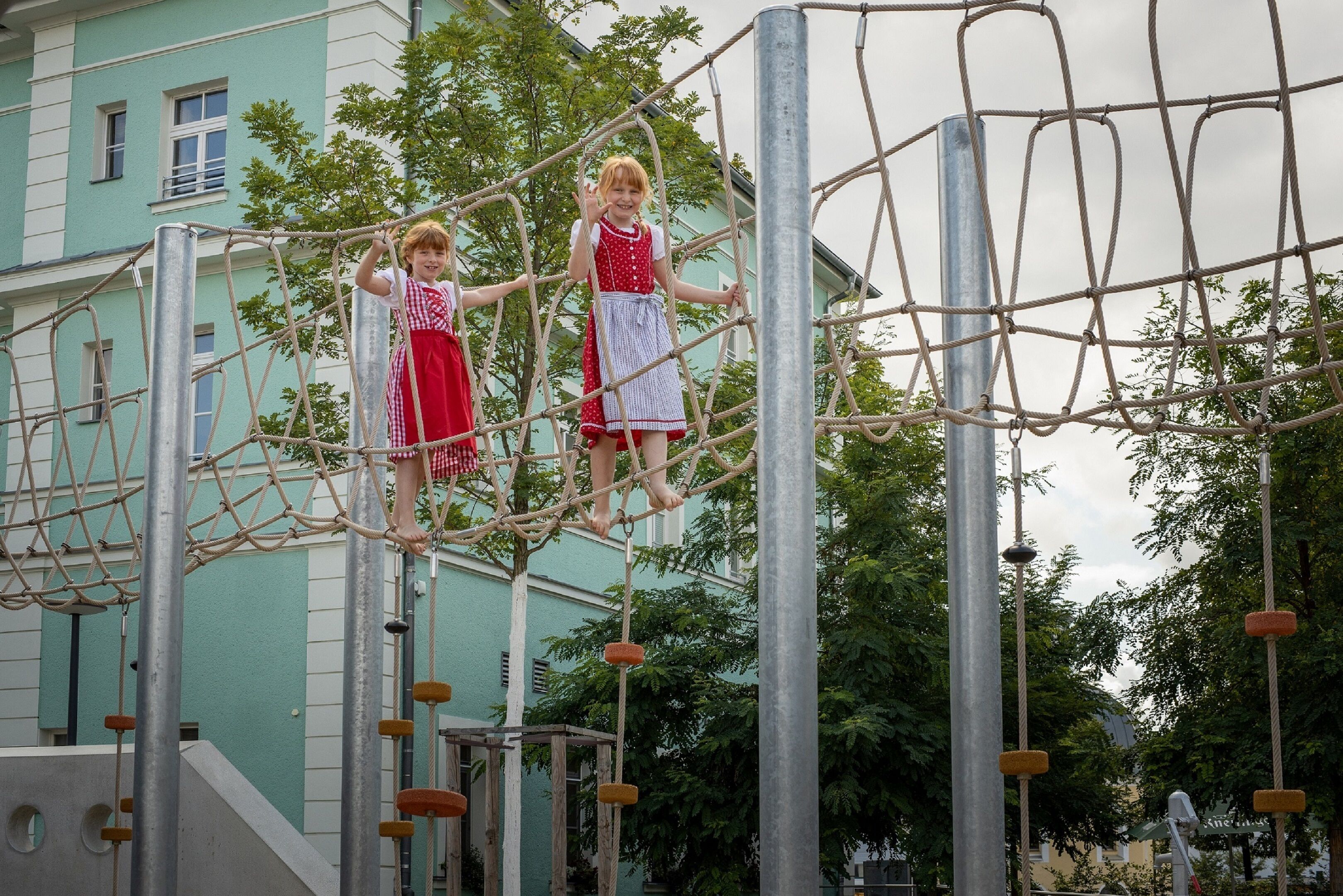 Zwei Kinder überqueren eine Seilbrücke in einem Spielbereich im Freien.