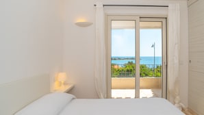 Un dormitorio con cama, una mesita de noche con lámpara, una puerta corrediza de cristal a un balcón y vista al mar.