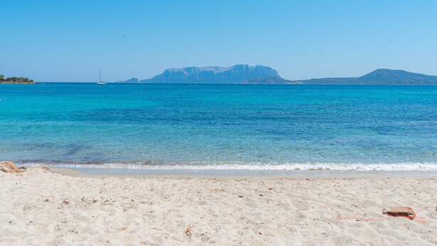 Una playa con aguas turquesas cristalinas, un velero y montañas a lo lejos.