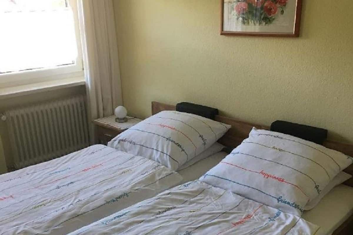 Una cama doble con ropa de cama blanca y varias almohadas, una mesita de noche con una lámpara y un cuadro de flores enmarcado en la pared.